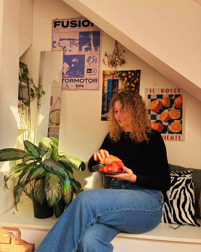 Woman on a built-in bench holding a plate with croissants, plants and posters behind her.